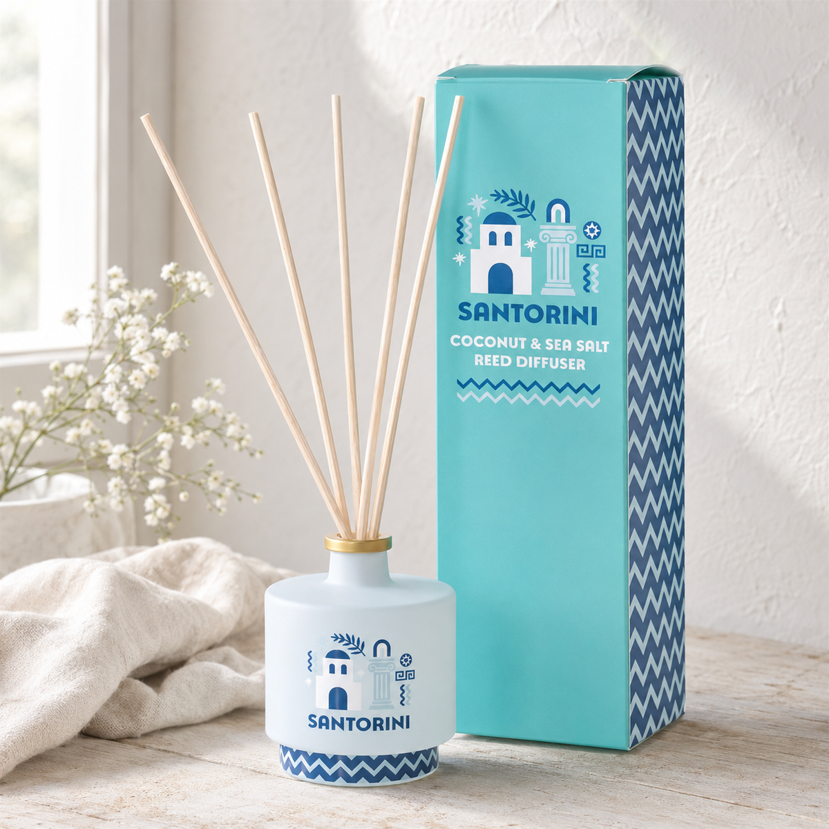 A Santorini coconut and sea salt reed diffuser bottle with natural reed sticks, shown next to its turquoise and blue packaging box on a light wooden surface.