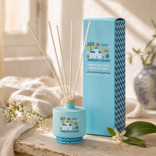 A light blue reed diffuser bottle with several natural reed sticks and a matching tall box, both printed with a tropical beach illustration and the text 'AMALFI COAST' and 'LEMON BLOSSOM REED DIFFUSER.' The scene is styled on a stone surface with soft fabric, flowers, and a vase in a sunlit interior.