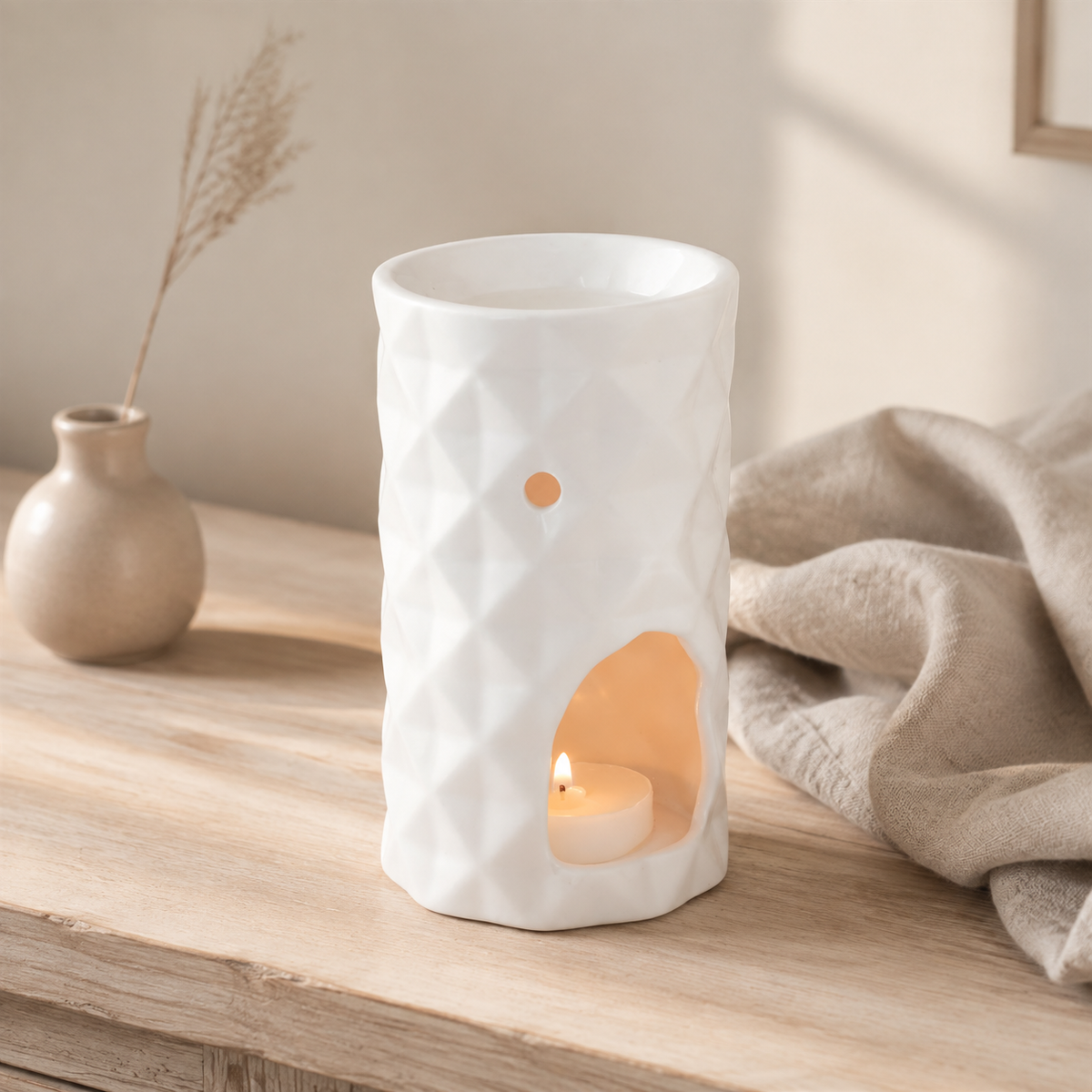 A white textured ceramic candle warmer with a geometric diamond pattern, featuring a cutout on the front that holds a lit tea light candle. The warmer is styled on a light wooden table next to a beige throw blanket and a small ceramic vase with dried grass.