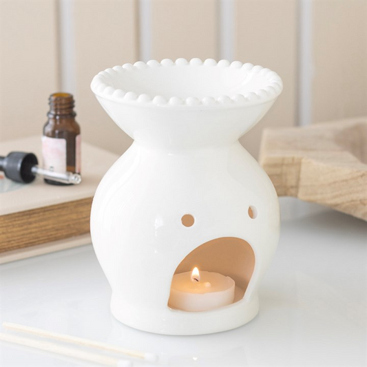 White ceramic oil burner with a lit candle inside on a surface.